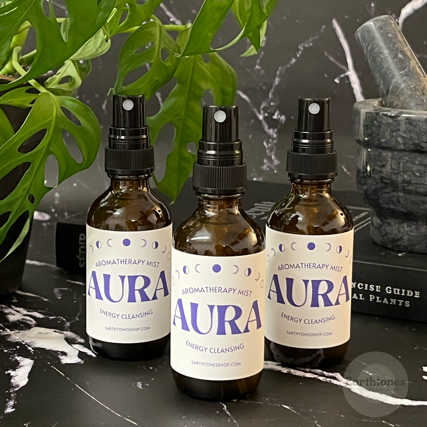 Three bottles of Aura aromatherapy mist on a dark surface with plants and stones in the background.