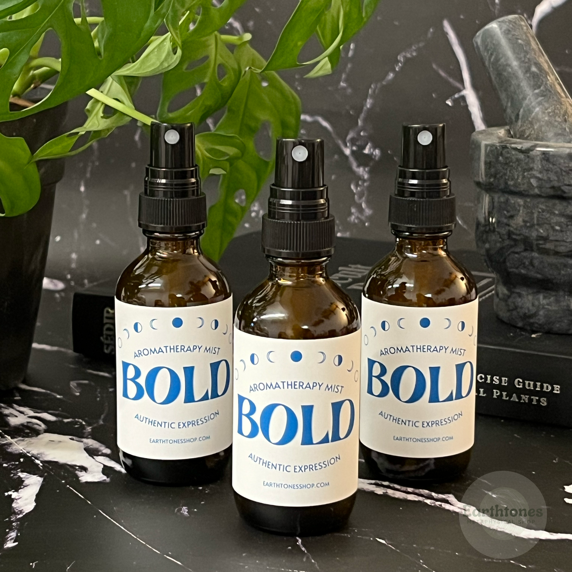 Three bottles of Bold Aromatherapy Mist on a dark surface with plants in the background.