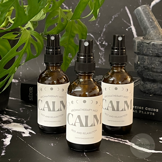 Three bottles of 'CALM' aromatherapy mist on a dark surface with plants and books in the background.