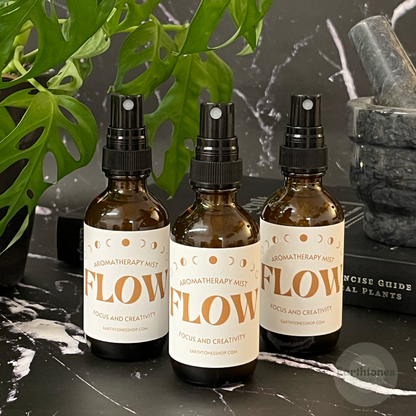 Three bottles of 'FLOW' aromatherapy mist on a dark surface with plants and books in the background.
