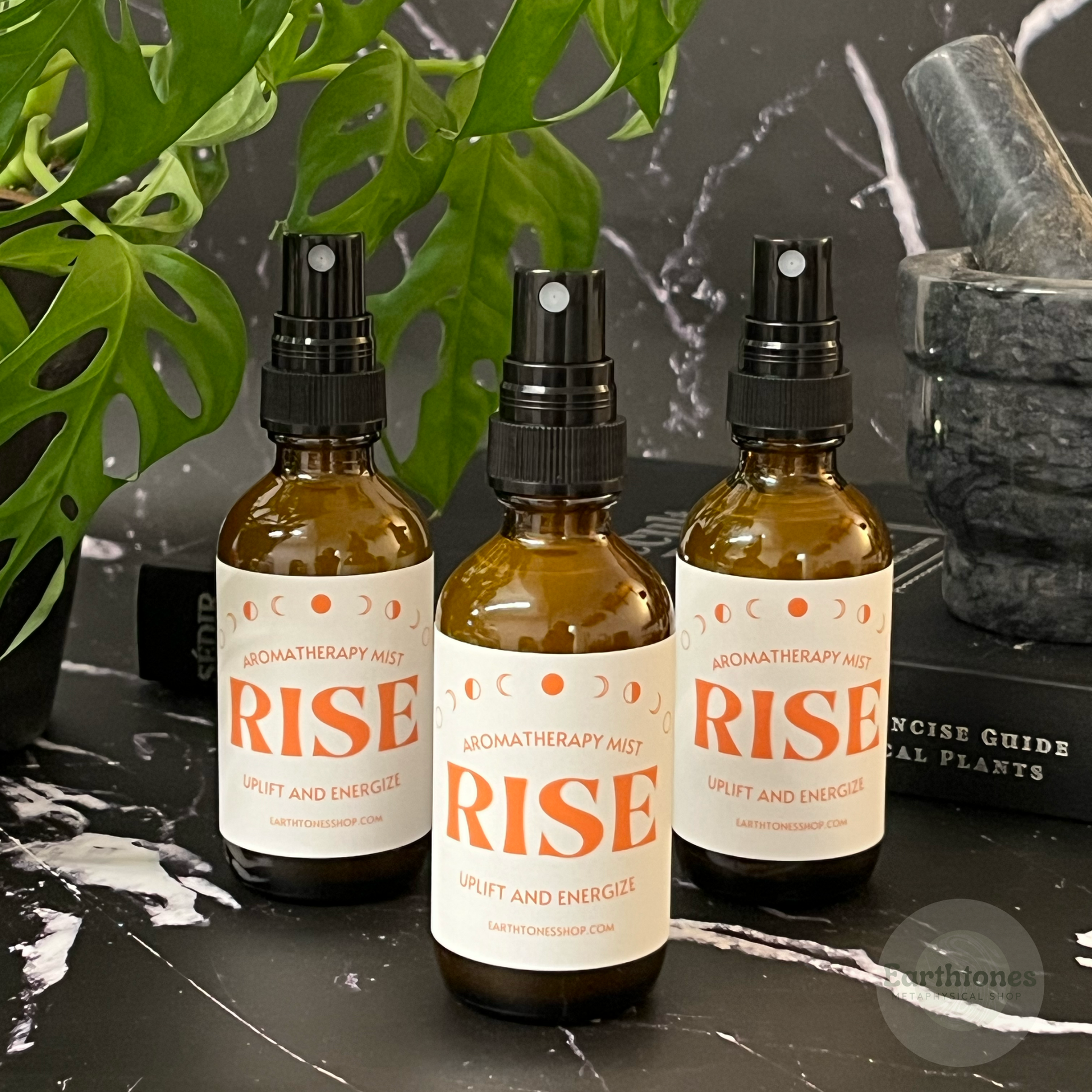 Three 'RISE' aromatherapy mist bottles on a dark surface with plants and rocks in the background.